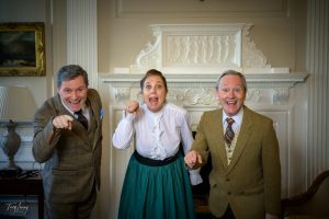 Three adults in vintage clothing stand indoors, excitedly pointing towards the camera and smiling. Behind them is an ornate white mantelpiece with classical reliefs, creating a lively scene that captures the joy of photography. The mood is animated and cheerful.