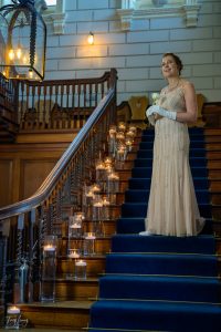 A woman in a vintage-style beaded gown and gloves stands on a grand wooden staircase lined with lit candles in glass holders, holding a white flower and smiling. The elegant, historic setting creates a timeless moment of photography.