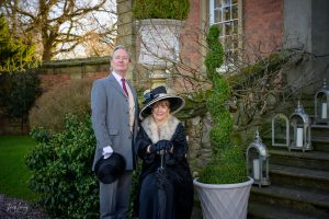 A man in a grey suit and a woman in a black dress with a fur collar and hat pose for photography beside a spiral topiary plant, in front of a brick building and stone steps adorned with lanterns.