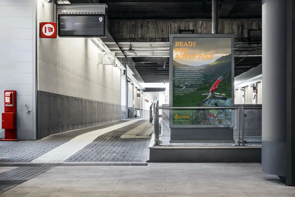 A bus station with empty platforms and a large advertisement reading READY FOR ADVENTURE, showing a person climbing a mountain. Photography accents the scene, with a fire extinguisher and emergency sign mounted on the left wall.