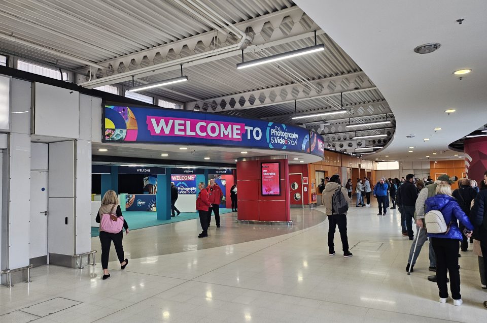 A spacious indoor venue with people walking and a large sign overhead reading WELCOME TO Photography & Video Show. The bright area, featuring white floors and ceiling, includes several information stands focused on photography.