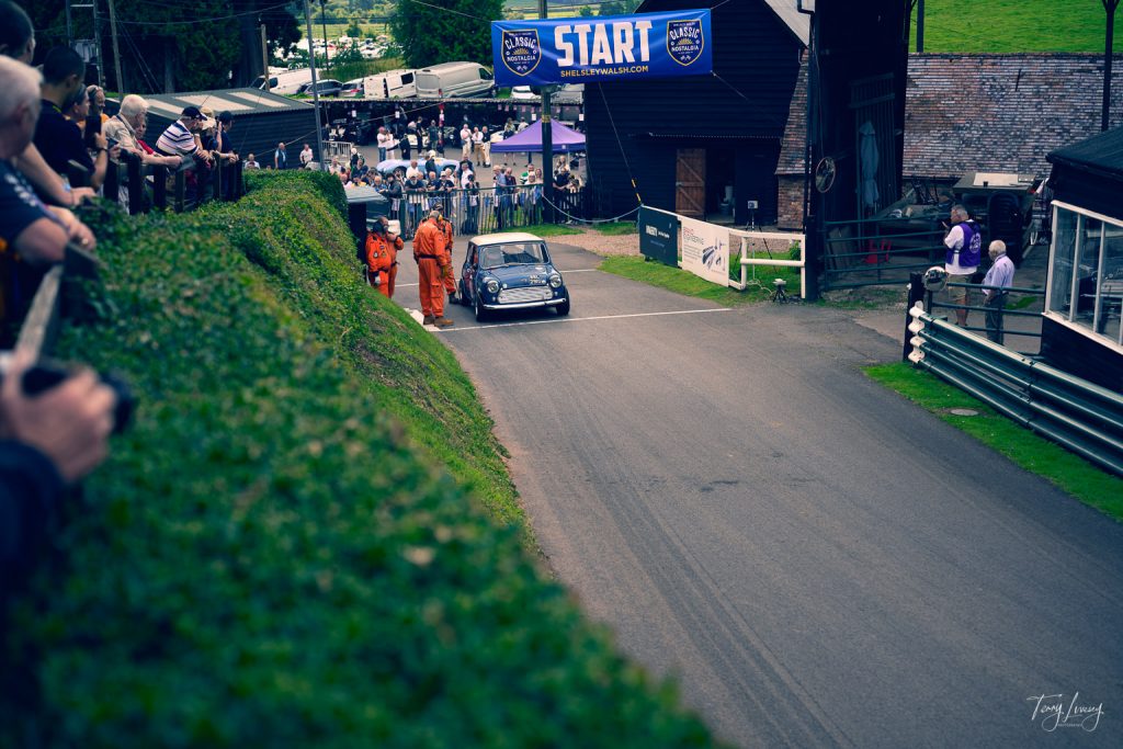 A classic black car approaches the start line of a hill climb race, captured in striking photography. Crowds watch as officials in orange suits stand nearby, framed by banners reading START above the road.