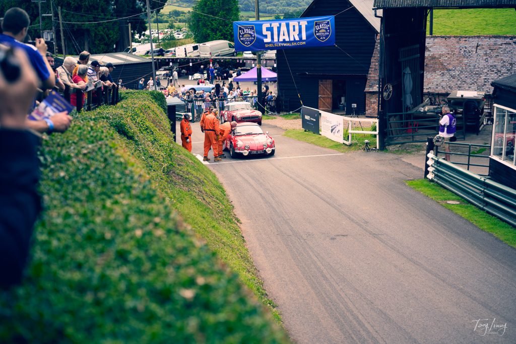 A classic red car is positioned at the start line of a hill climb race, captured in stunning photography, with race officials in orange suits nearby and spectators watching from both sides. A blue START banner is displayed overhead.