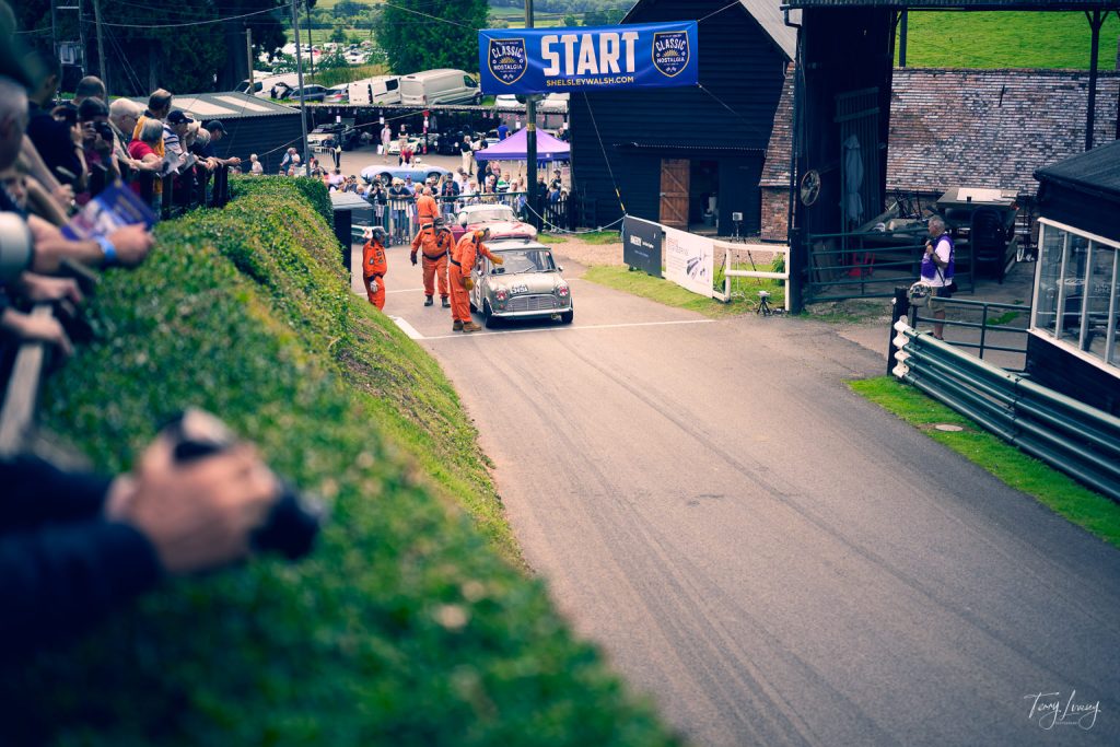 A vintage car approaches the starting line at a motorsport event, guided by several people in orange suits. Spectators line a hedge, watching and snapping photographs as a large START banner hangs above the track.