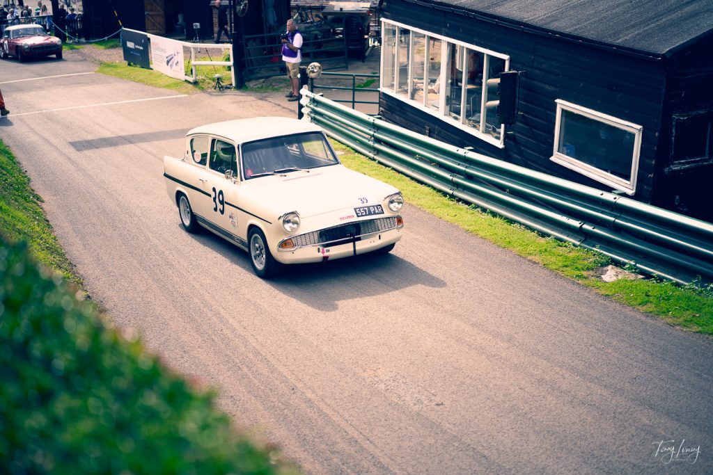 A vintage white racing car with the number 39 on the door drives on a tarmacked track by a black building, green crash barriers, and grassy areas during a motorsport event, captured in stunning photography.