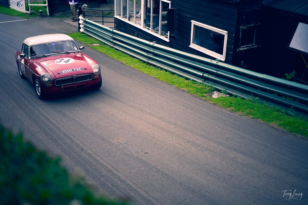 A classic red racing car with the number 34 on the bonnet speeds along a curving tarmac road next to a black building and green metal crash barrier. The photography captures the energy of a vintage car race or rally event.
