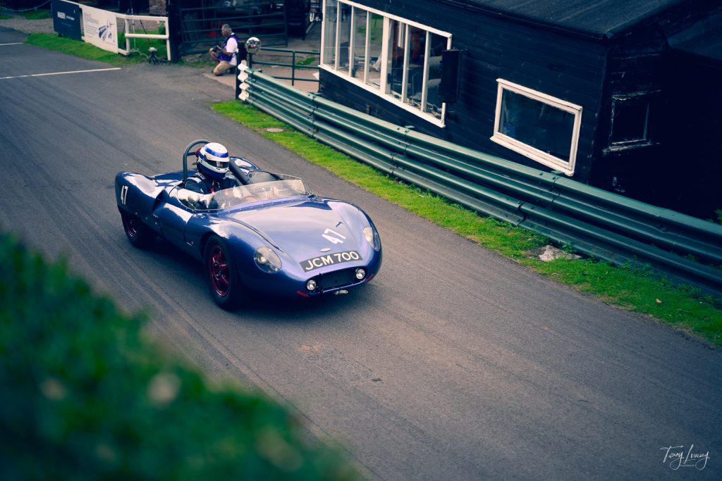 A blue vintage racing car with the number 17 and registration plate JCM 700 is captured in striking photography, driven by a helmeted driver on a tarmac track beside a black wooden building at a motorsport event.