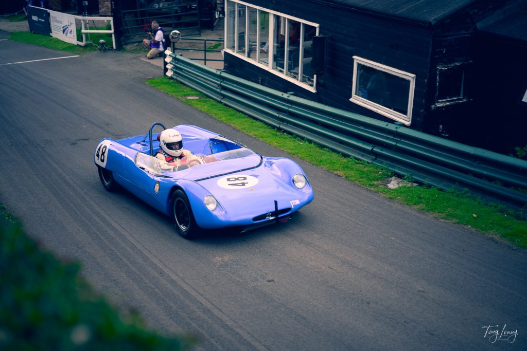 A blue vintage racing car with the number 48 on the bonnet is captured in striking photography as a helmeted driver speeds along a tarmac race track, passing a black building with large windows and green railings.