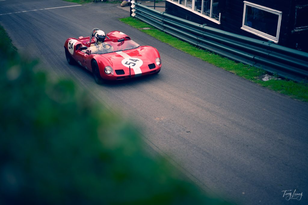A vintage red racing car with the number 54 on its bonnet speeds round a corner on a tarmac track, captured in stunning photography by a helmeted driver. Green barriers and a building are visible along the side of the track.
