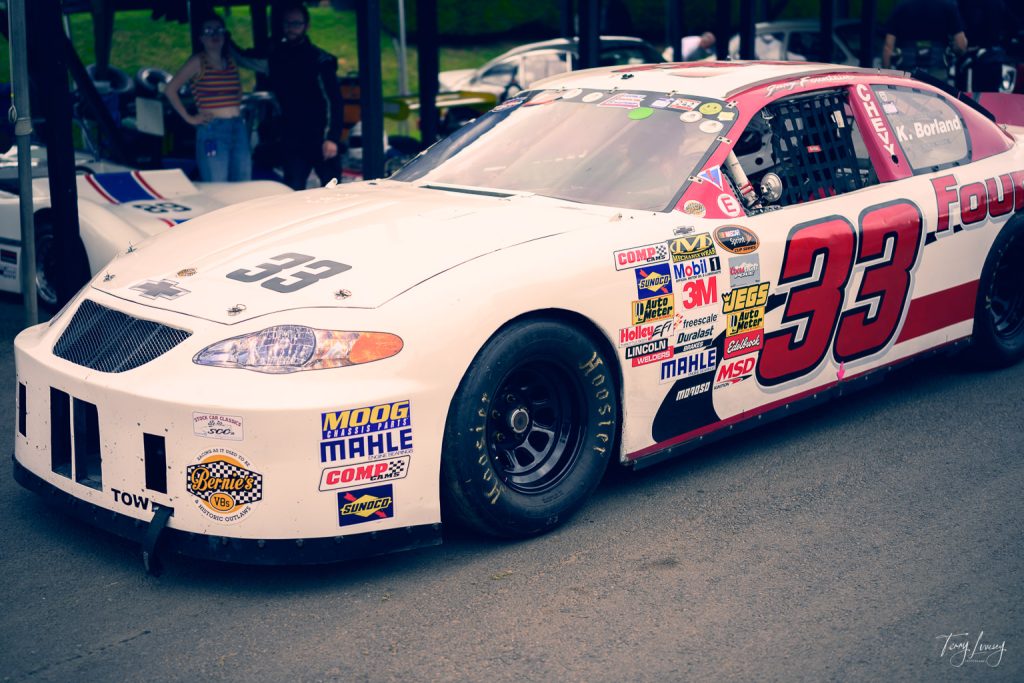 A white and red stock race car numbered 33 is parked in a paddock area, covered with sponsor stickers; people and other vehicles are visible in the background, capturing a dynamic moment in motorsport photography.