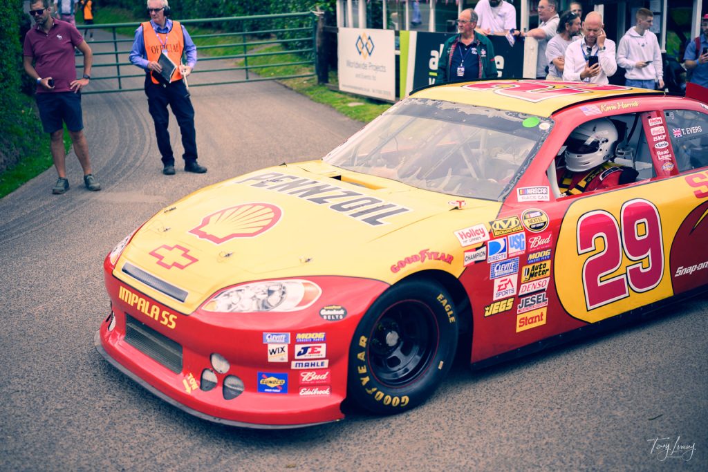 A striking example of motorsport photography, this image captures a yellow and red racing car with the number 29 and Shell Pennzoil branding parked on a road, surrounded by event staff and spectators as the helmeted driver waits inside.