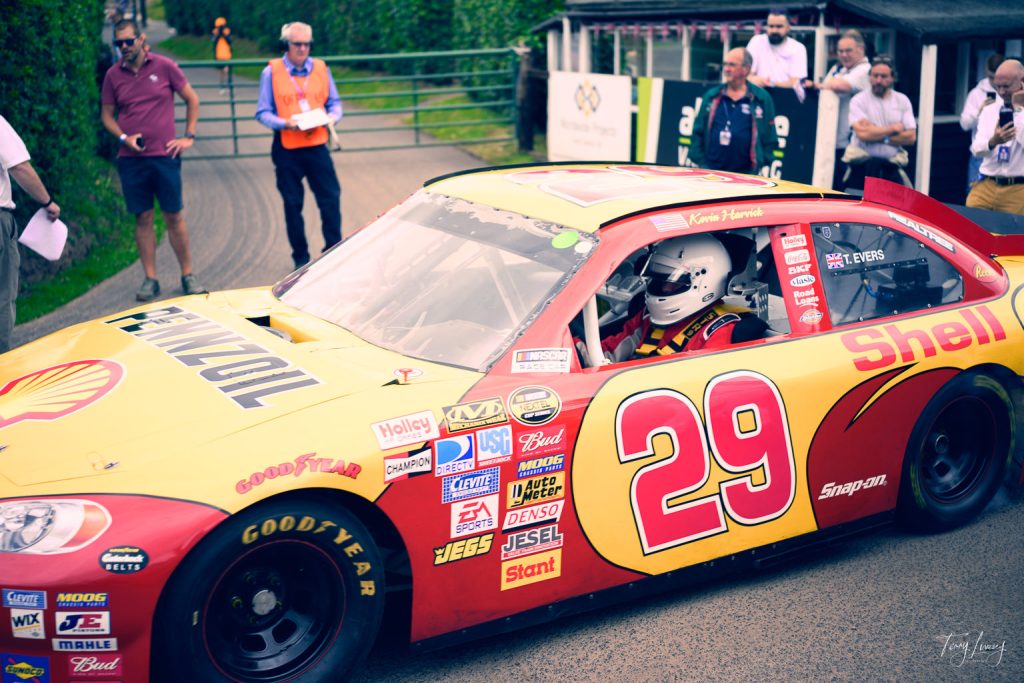 A yellow and red number 29 Shell-sponsored racing car with a helmeted driver is parked, as several people—some in orange vests and face masks—stand nearby in an outdoor setting, captured in vivid detail through photography.