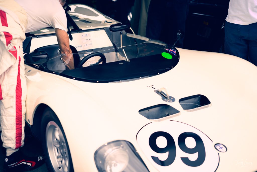 A person in a white racing suit is photographed leaning into a vintage white racing car with the number 99 on the bonnet, preparing or inspecting the interior.
