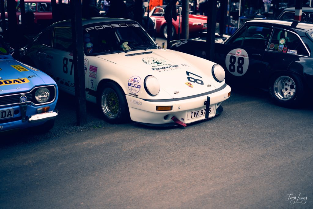 A white Porsche racing car with the number 67 on the bonnet is parked between two classic racing cars in a shaded area, captured in striking photography at a car show or racing event.