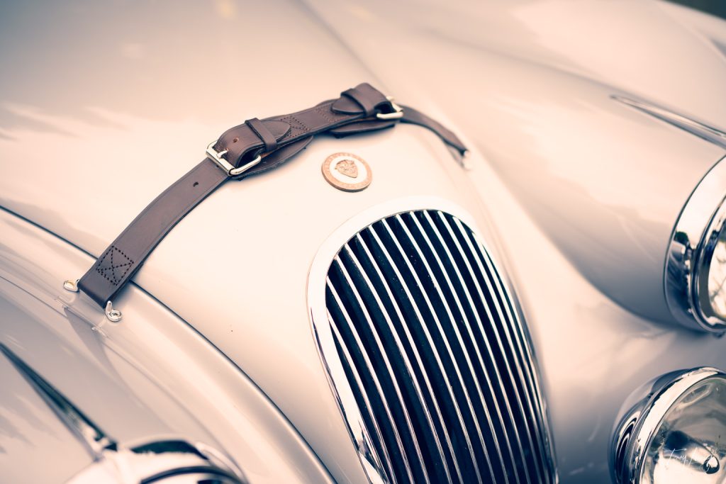 Close-up photography of a vintage car’s front bonnet shows a leather strap, classic grille, and two headlamps, with a round badge centred above the grille. The image has a soft, retro tone.