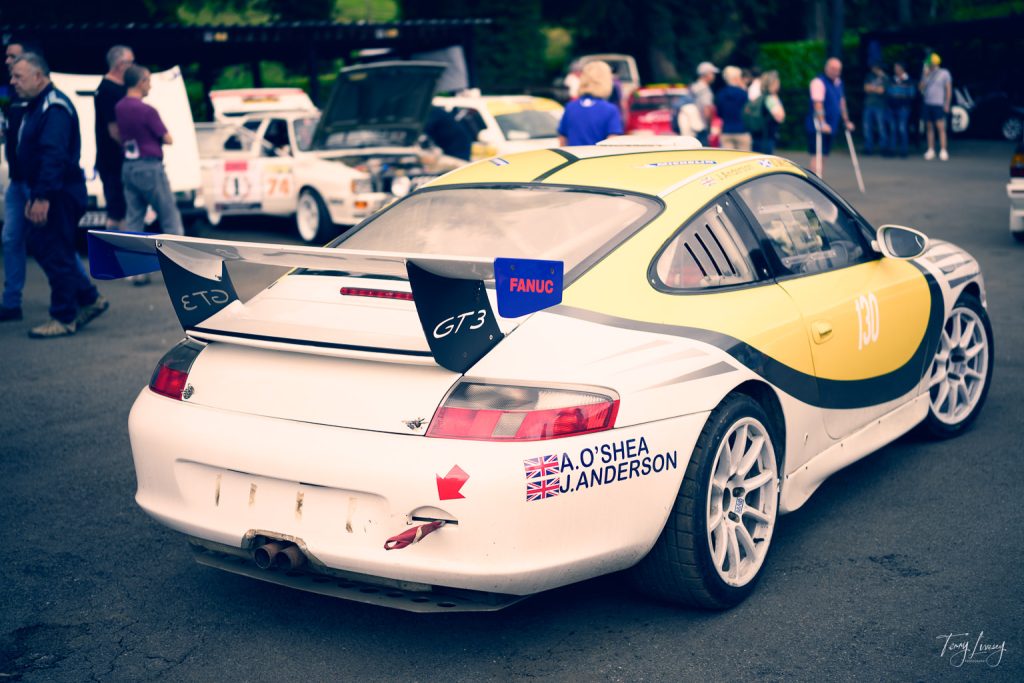 A white and yellow Porsche GT3 racing car with sponsor decals is parked at a car event, surrounded by people and other classic cars in the background, making it a striking subject for motoring photography.