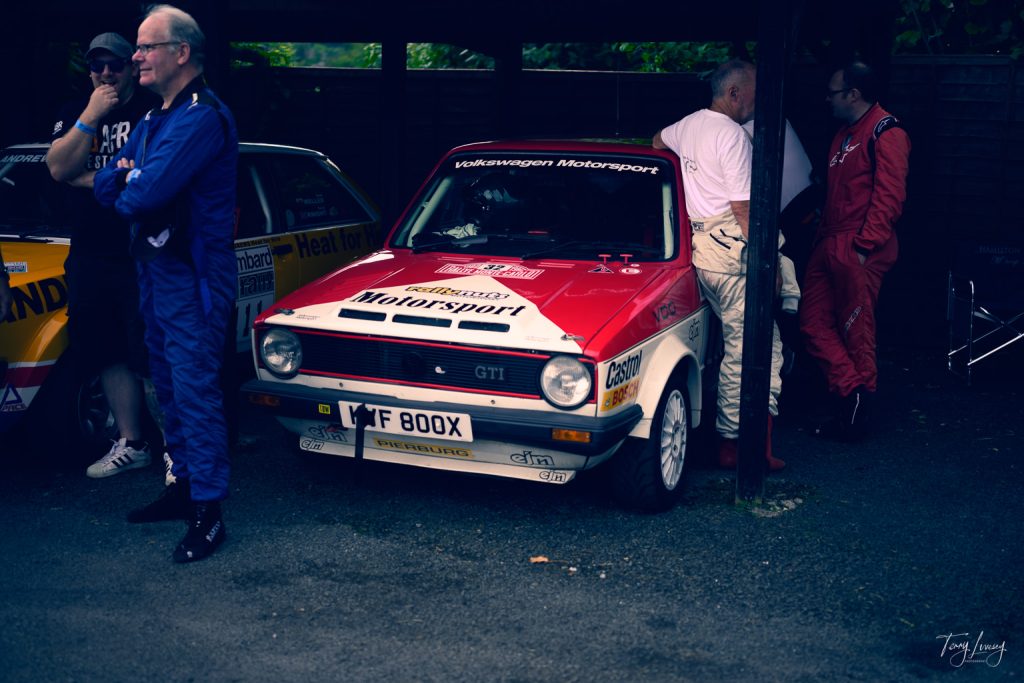 A vintage Volkswagen Motorsport GTI rally car is parked under a shelter in this striking photograph. Four men in racing suits stand and converse around the car, while another yellow rally car is partially visible to the side.