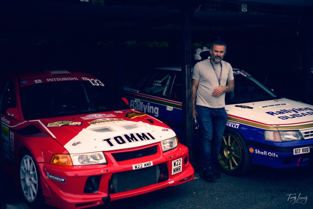 A man stands between a red Mitsubishi rally car with TOMMI on the bonnet and a white Subaru rally car with blue and yellow accents in a dimly lit garage, capturing a moment of automotive photography.