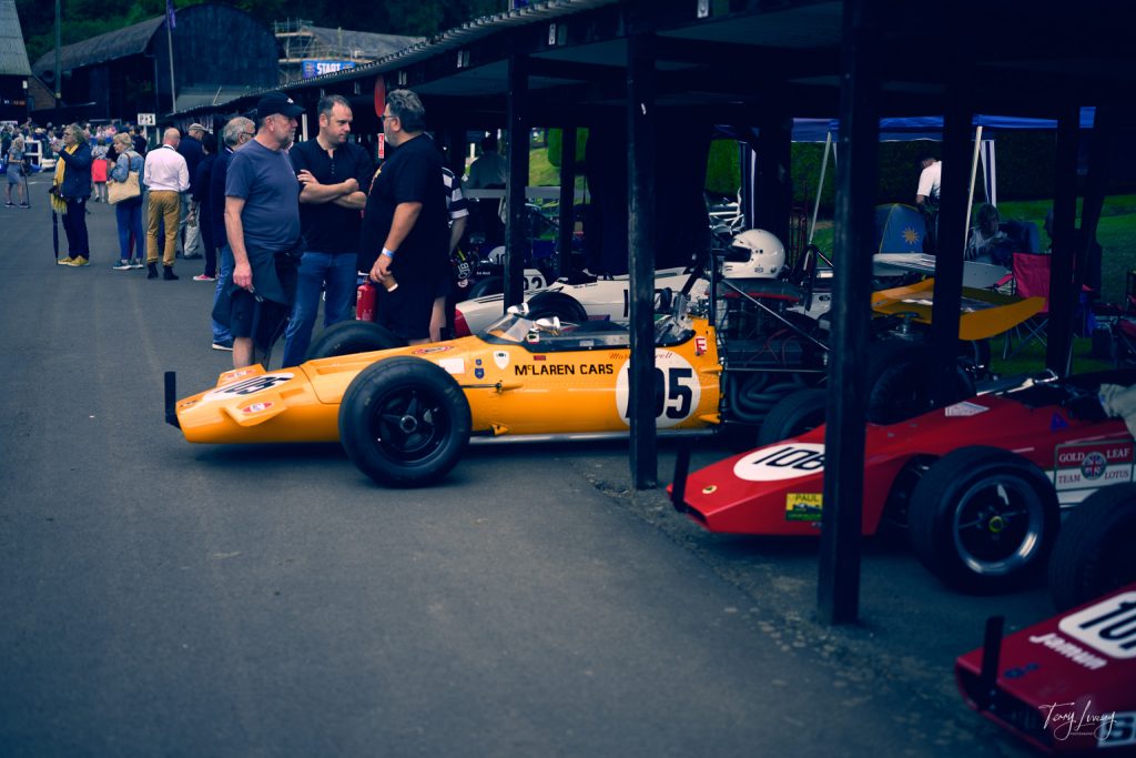 People stand and talk beside a classic yellow McLaren racing car parked under a shelter, while nearby drivers in helmets pose for photographs with other vintage racing cars at a motorsport event.