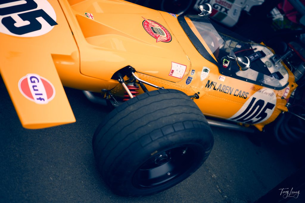 A close-up angled photography shot of a vintage orange McLaren racing car, numbered 105, highlights its large rear tyre, decals, and open cockpit at what appears to be a motorsport event or car show.