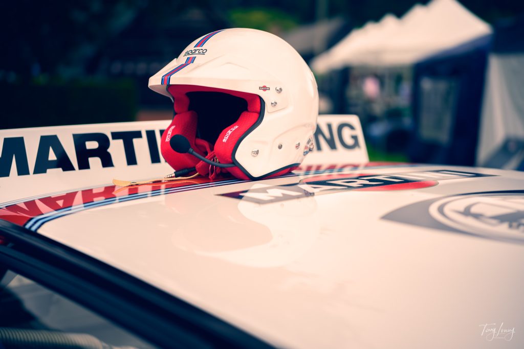 A white racing helmet with a microphone rests on top of a “Martini” branded car with racing stripes—captured in striking photography, with white marquees and blurred greenery visible in the background.