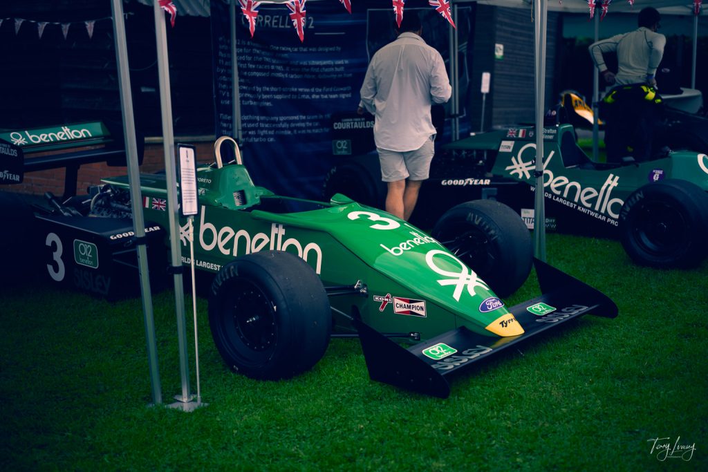 A green Benetton Formula 1 racing car with the number 3 is displayed outdoors under a canopy in this captivating photograph. A man in light clothing stands nearby, with another similar racing car partially visible next to it.