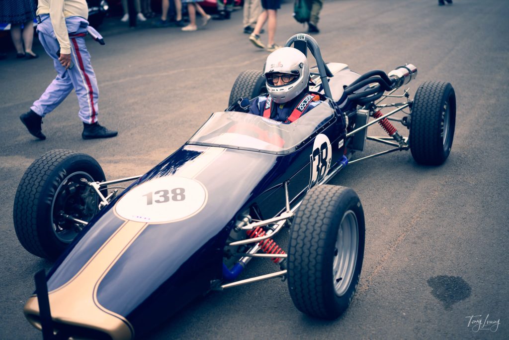 A person wearing a helmet and racing suit sits in a vintage blue and gold open-wheel racing car marked with the number 138, parked on a tarmac track. This striking moment of motorsport photography also captures people standing in the background.