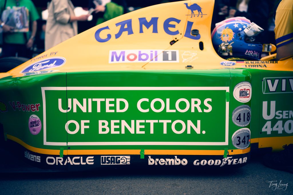 Close-up photography of a yellow and green Formula 1 car sidepod, featuring large green United Colours of Benetton branding, sponsor logos, part of the rear tyre, and a blurred crowd in the background.