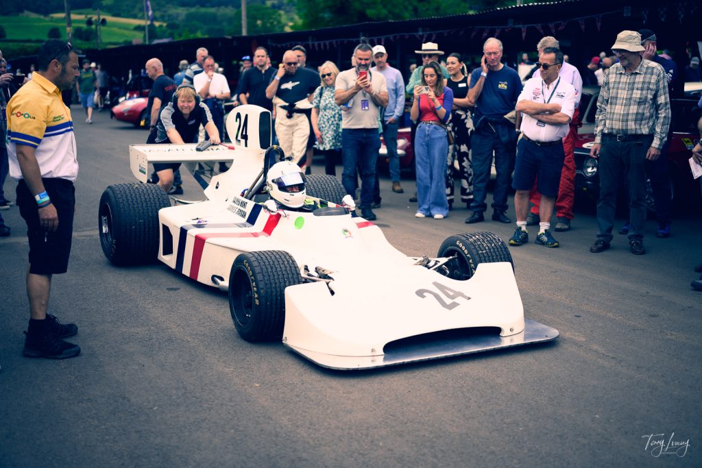 A vintage white racing car with the number 24 is driven by a helmeted driver, captured in striking photography, surrounded by a crowd of spectators and officials at a motorsport event.