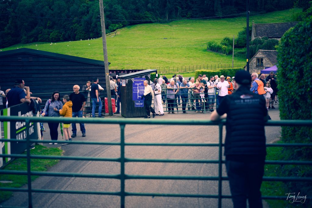 A crowd gathers near a green metal gate at an outdoor event in a rural area with grassy hills and trees, captured through photography. One person stands in the foreground, leaning on the gate and observing the scene.