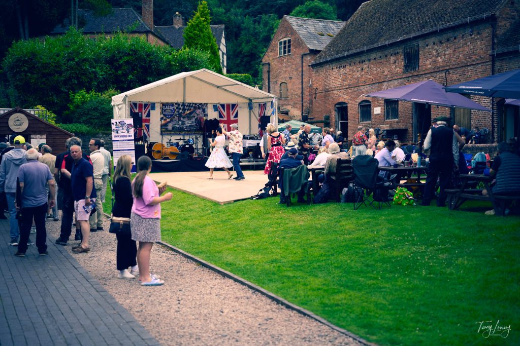 Outdoor event photography captures people seated at picnic tables, watching dancers perform on a small stage adorned with Union Jack flags. Historic brick buildings and green lawns surround the lively scene.