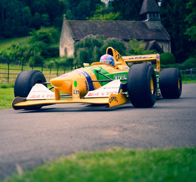 A yellow and green Formula 1 racing car speeds along a road flanked by lush greenery and an old stone building with a steeple, captured in stunning photography. Sponsor logos and a helmeted driver complete the dynamic scene.