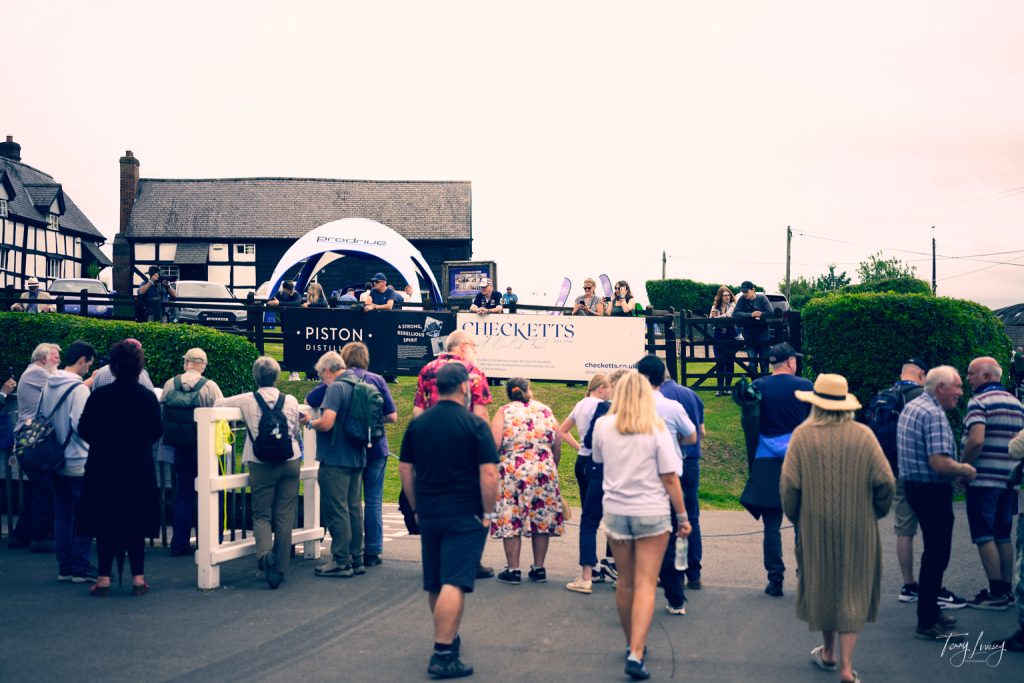 A crowd of people stands outside a fenced area, watching an outdoor event with a stage, banners, and a canopy. Some wear hats and jackets; traditional buildings provide a picturesque backdrop for photography enthusiasts.