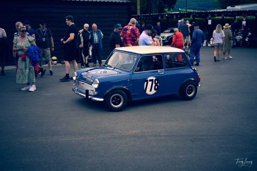 A classic blue Mini Cooper with the number 178 on its door is parked on a street, captured in vibrant photography, surrounded by people in vintage-style clothing at an outdoor event.