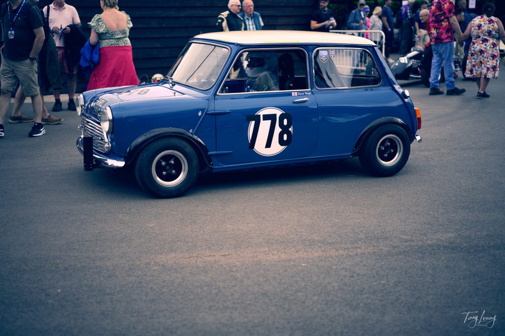 A blue classic Mini Cooper racing car with the number 178 on its door is parked on a street; this vibrant scene is captured in stunning photography, with people walking and standing in the background.