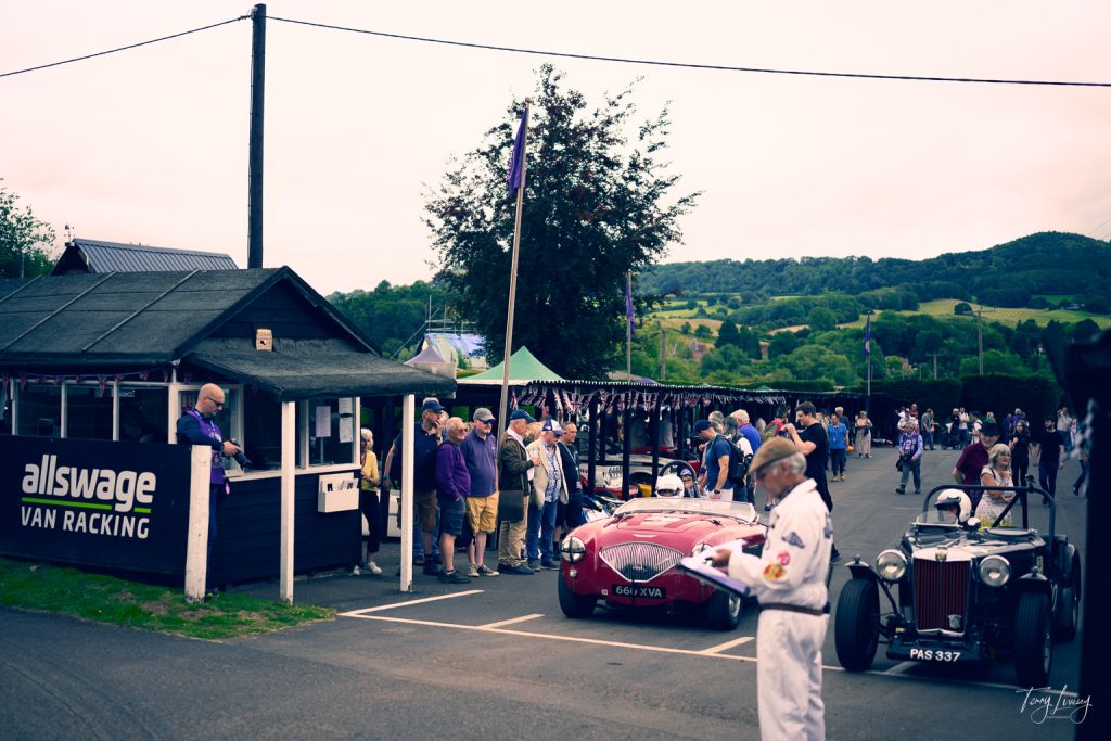 A crowd gathers near a small wooden building labelled allswage VAN RACKING, with classic cars and a man in vintage racing attire in the foreground, set against a backdrop of green hills and overcast skies.