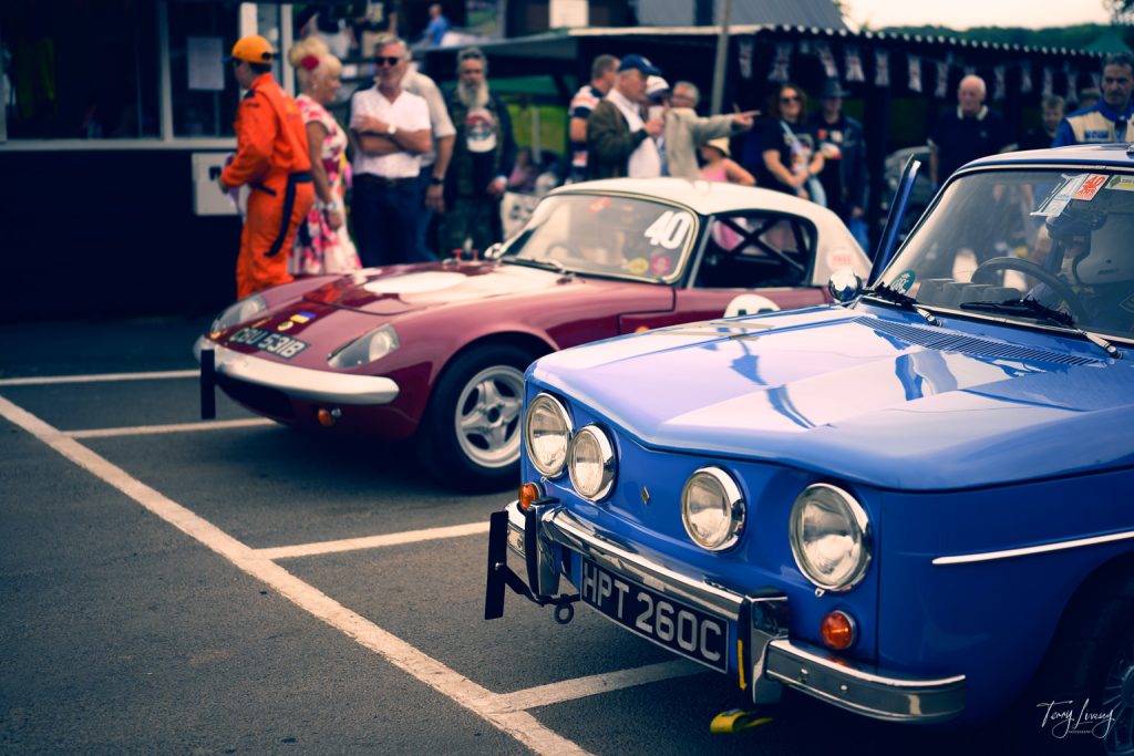 Two classic cars, one blue and one red, are parked at a car show. People stand nearby, some in orange overalls. The scene is lively, with spectators admiring the vehicles and socialising.