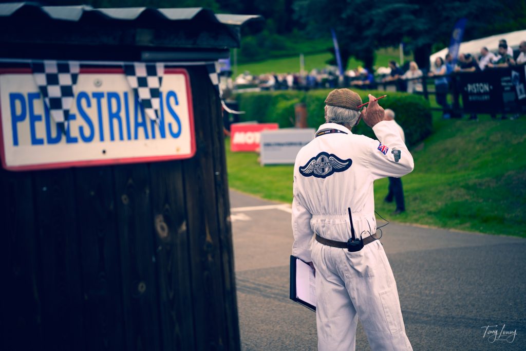An older man in a white jumpsuit and cap holds a clipboard and points, standing near a Pedestrians sign at an outdoor event with a crowd in the background.