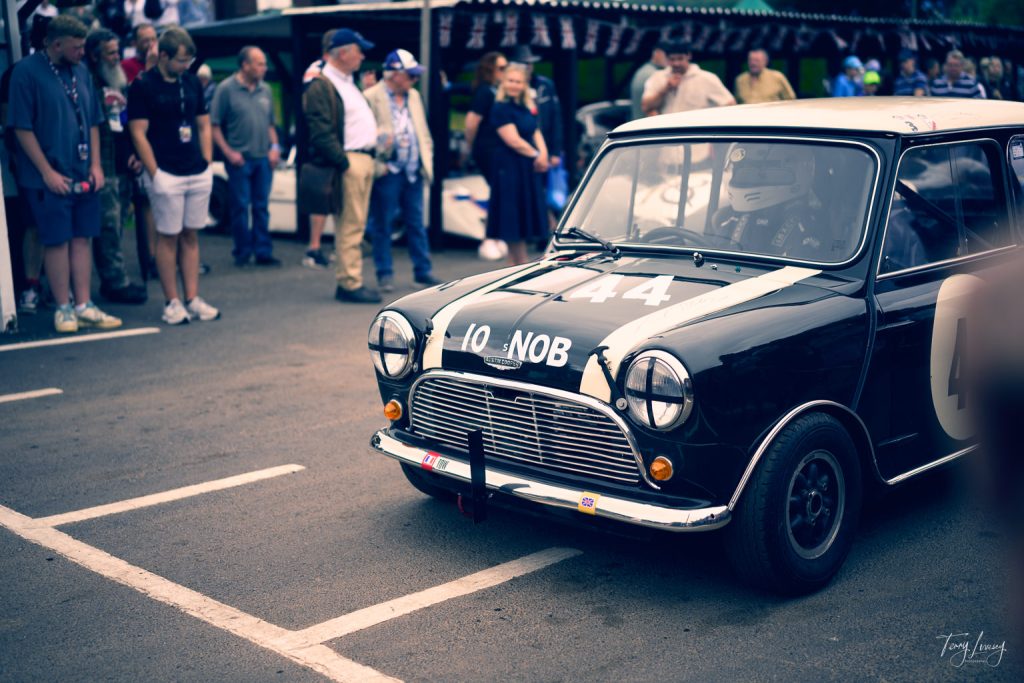 A classic black Mini Cooper car with racing numbers is parked on a road, surrounded by a group of people watching at an outdoor event. The driver, wearing a helmet, is visible through the windscreen.