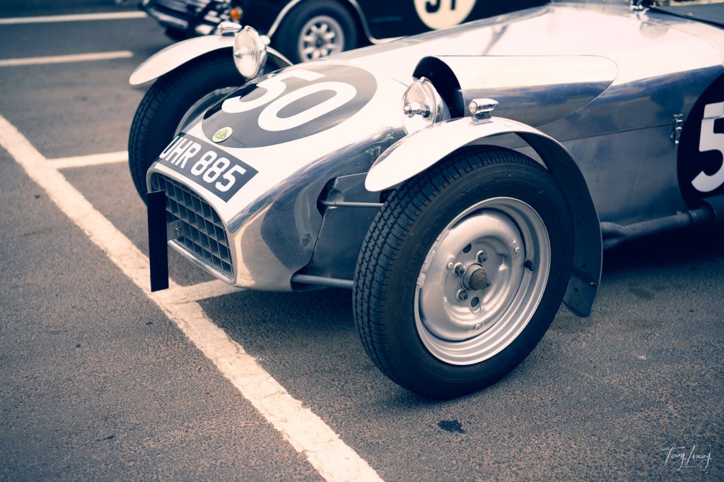 A vintage silver racing car with the number 50 on the bonnet is parked in a car park, featuring classic round headlamps, wire wheels, and a visible number plate reading “XMR 885”.