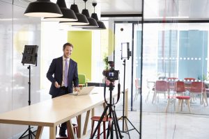 A man in a suit stands at a table with a laptop, smiling whilst being filmed in a modern office. Camera equipment and studio lights, essential for professional photography, surround him, whilst red chairs are visible in the background.