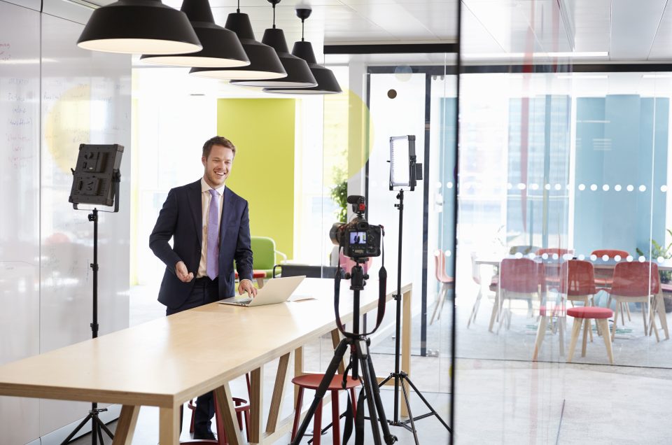 A man in a suit stands at a table with a laptop, smiling whilst being filmed in a modern office. Camera equipment and studio lights, essential for professional photography, surround him, whilst red chairs are visible in the background.