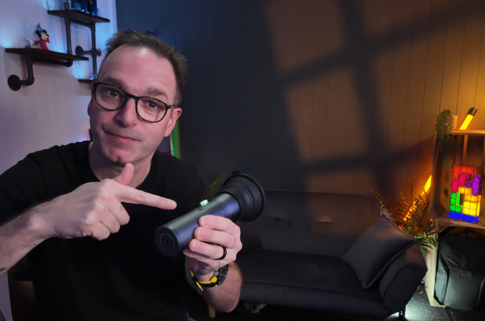 A man with glasses and a tattoo on his arm points to a torch he is holding, sitting in a cosy room with a sofa, shelves, and colourful lights in the background.