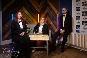 Three people in suits sit at a table with plants, against a colorful wall. "The WiNN Show" logo visible.