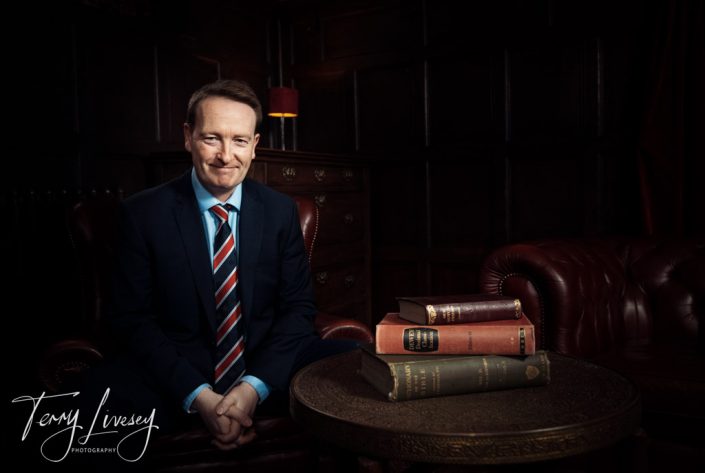 OSH - Headshot - Paul Kilbride sits in a dimly lit room with leather chairs and stacked antique books nearby.