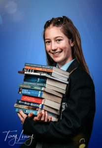 OSH holds a stack of books including 'The Boy in the Striped Pyjamas' and 'A Monster Calls', wearing a school uniform.