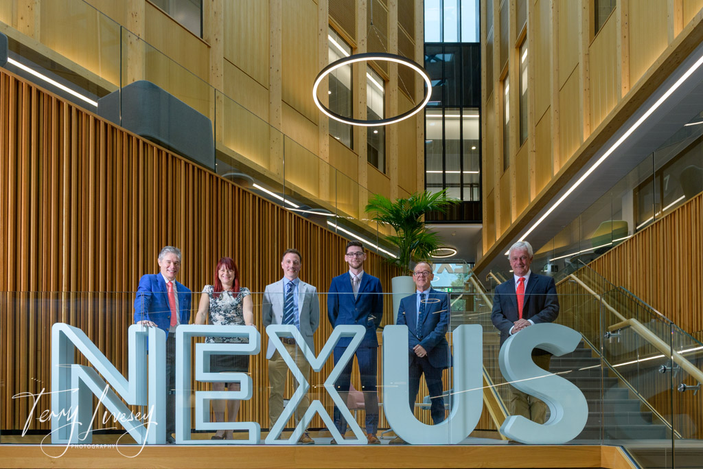 explic8 - Group of six individuals stand behind large 'NEXUS' sign inside a modern building with wooden walls.