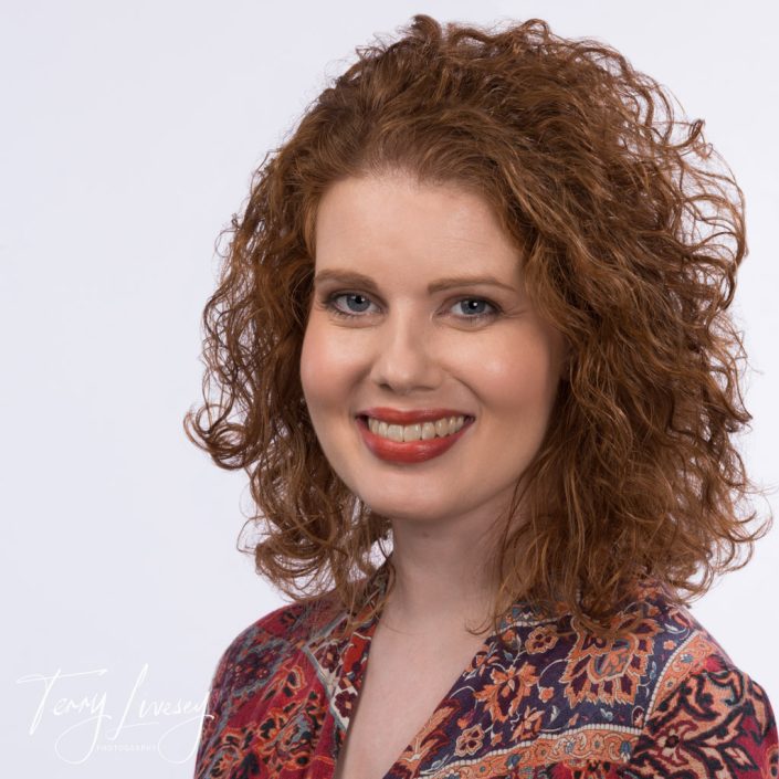 Charlotte Scriven smiling in a headshot with curly hair and patterned attire against a plain background. Professional appearance.