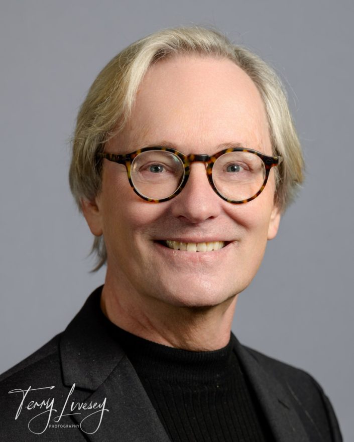 BMTC Headshots - A man with glasses smiles while posing for a professional portrait in front of a gray background.