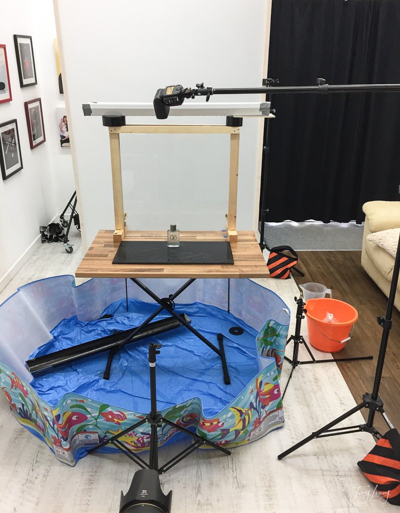 BTS shot showing a glass panel with an aftershave bottle in front. All on a table which is in a paddling pool to collect the water that will be poured over it.